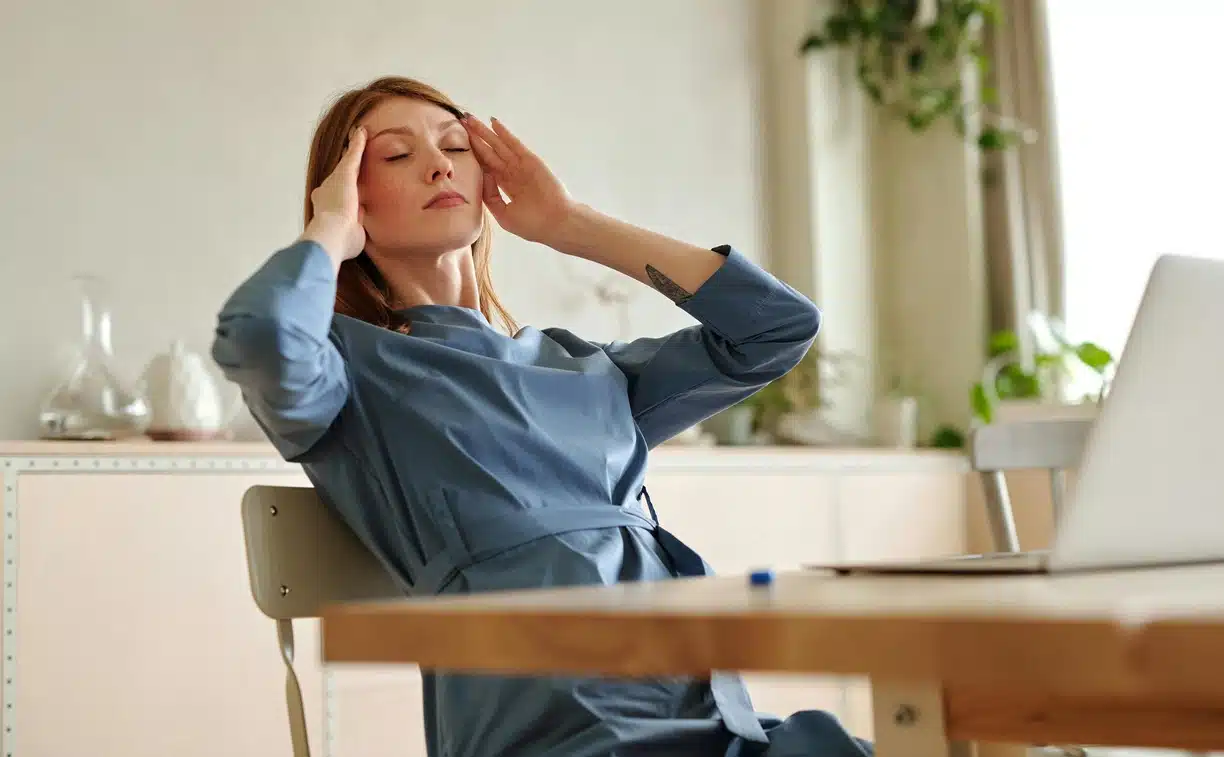 Woman sitting at a table with a laptop, woman is holding both sides of her head because of a migraine